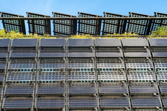 View of solar panels installed on a building wall and roof with green vegetation visible against a clear blue sky in The Netherlands.