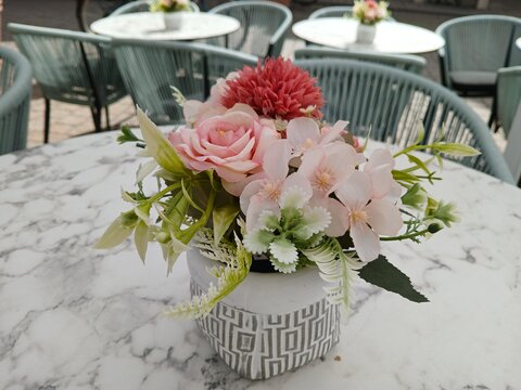 A delicate pink floral arrangement rests on a marble cafe table, creating a romantic summer atmosphere among elegant outdoor seating and greenery.