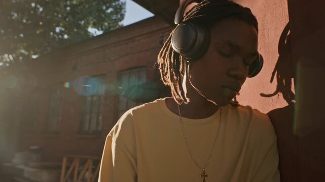 African American teenager wearing headphones leaning against wall outdoors, listening to music with closed eyes in slow motion. Clip is ideal for audio technology ads and music streaming promos