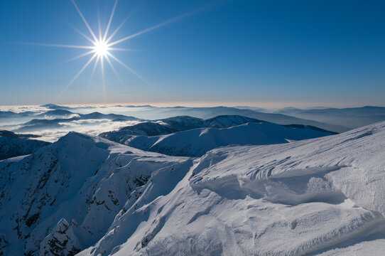 View of the snow-covered peak of Dumbier with a bright sunburst in a clear blue sky and rolling mountain ranges above a sea of clouds, Nizke Tatry, Zilina Region, Slovakia.