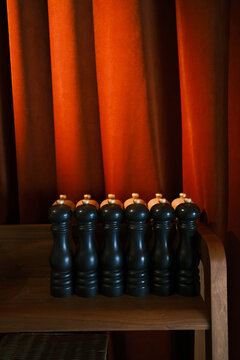 Rows of black pepper grinders arranged on a wooden restaurant station against a warm red curtain, illustrating hospitality preparation, organization and service readiness.