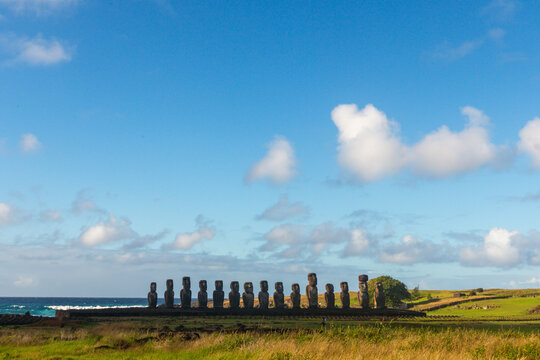 View of the iconic Moai statues standing in a row on the coast of Ahu Tongariki with grassy fields and the blue ocean in Easter Island, Valparaiso, Chile.