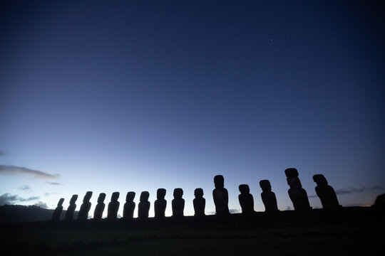 View of the silhouetted Moai statues standing in a row against a twilight sky at Ahu Tongariki on Easter Island, Valparaiso, Chile.