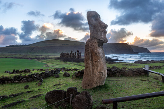 View of the iconic Moai statues at Ahu Tongariki on Easter Island, set against a dramatic sunset sky with rolling green hills and the rocky coastline of Easter Island, Valparaiso, Chile.