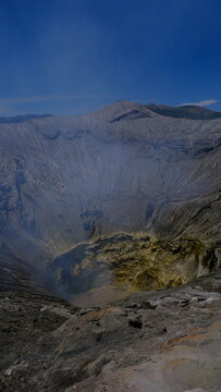 View of the volcanic crater of Mount Bromo with rugged rocky slopes, sulfurous yellow deposits, and rising smoke under a clear blue sky in East Java, Indonesia.