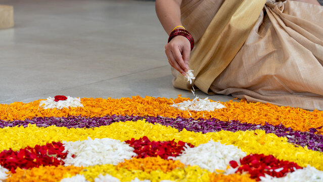 View of a woman in a traditional saree creating a colorful pookalam flower rangoli for the Onam festival, placing white flower petals onto the intricate floral pattern on the floor in India.