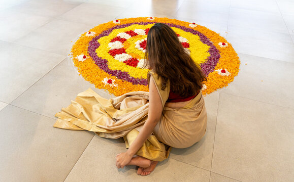 View of a woman in a traditional sari sitting on a tiled floor while creating a colorful Pookalam floral arrangement for the Onam festival in India.