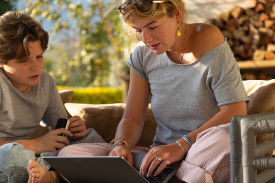 young woman using laptop computer on outdoor couch with younger brother looking on