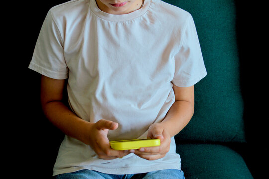 Young child in white t-shirt holding a yellow smartphone with both hands while sitting on a dark teal sofa in a dimly lit indoor setting
