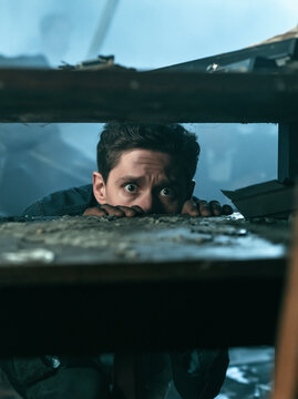 Scared young man hiding under wooden table looking out with wide terrified eyes