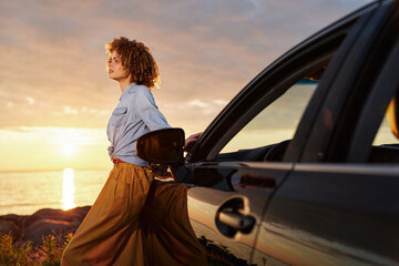 Naklejka premium A woman with curly hair in a white blouse and flowing skirt stands beside a car at sunset, capturing a carefree lifestyle on a coastal road and beach.