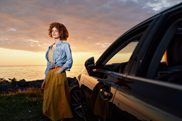 Naklejka premium Casual woman posing beside a car at sunset, wearing a denim shirt and yellow dress, capturing a relaxed lifestyle moment outdoors with warm light.