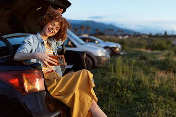 Fototapeta premium Smiling woman with curly hair sits on a car trunk taking a selfie. Outdoor parking lot beside a grassy field. Casual lifestyle, travel, wellness vibe and carefree mood.