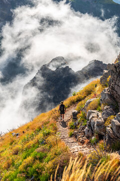 Hiker trekking along dramatic mountain stone path from Pico do Arieiro to Pico Ruivo in Madeira surrounded by dramatic rolling clouds and golden sunlight.