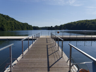 Fototapeta premium Visitors walk on a wooden pier by a river surrounded by trees on a sunny day in the countryside