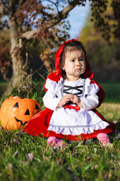 Toddler in Red Hooded Halloween Costume Sitting With Pumpkin Pail