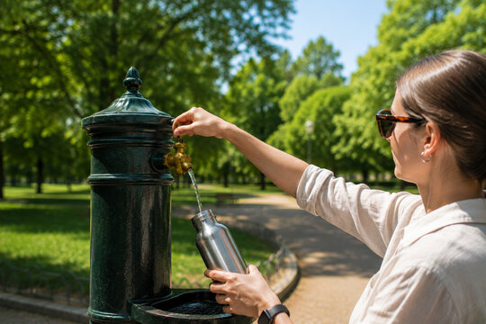 A man fills a reusable stainless steel bottle with water from a public drinking fountain in a European park on a hot summer day. Concepts: hydration, climate change adaptation, active lifestyle.
