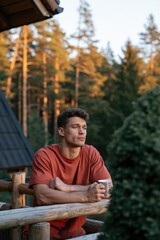 Fototapeta premium Young man on log cabin balcony holding a mug at sunset among pine trees for outdoor lifestyle background and travel banner