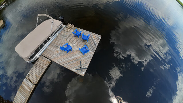 A wide angle overhead drone perspective looks down at a wooden lake dock detailed with blue adirondack chairs and a parked pontoon boat under a bright sky with clouds.
