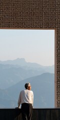 Naklejka premium Man standing on a balcony framed by ornate screen looking at misty mountain range for travel background and contemplative poster design
