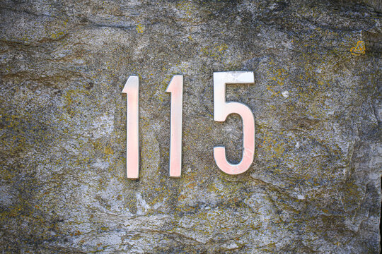 House number 115 sign in polished metal letters fixed on a lichen covered stone wall, displaying digit one one five in classic address numeral style