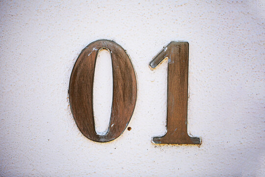 House number 01 sign crafted in weathered wood, mounted on a rough painted wall, showing rustic carved zero and one digits with vintage aged metallic trim outdoors