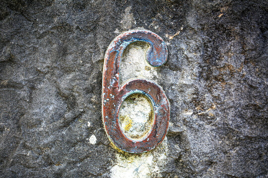 House number six sign in rusted metal, fixed onto a coarse stone wall, displaying a single weathered numeral with corroded patina and aged surface texture