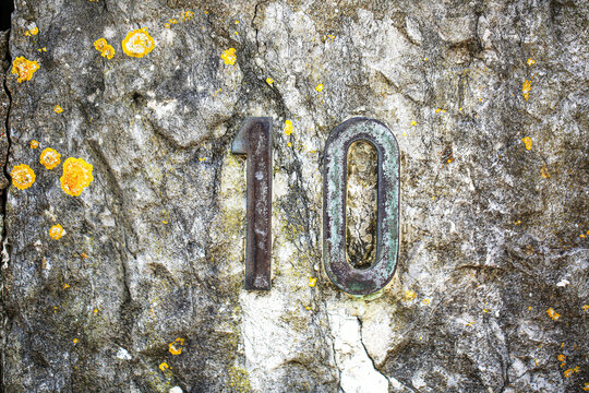 House number 10 sign in aged metal digits, mounted on a lichen covered stone wall, showing one and zero numerals with weathered rustic surface finish