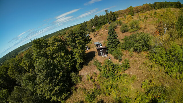 A high angle wide shot looks down over a small dark tiny house cabin nestled in a grassy clearing on a rugged woodland hill under a bright blue sky filled with clouds.