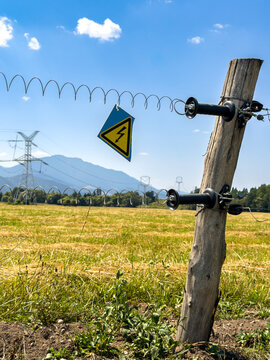 Wooden electric fence post with warning sign. A yellow high voltage placard hangs from coiled wire attached to a timber pole in a sunlit pasture.