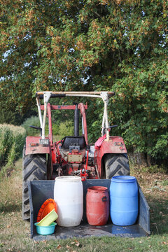 A red tractor transports blue, white, and red water barrels in a rear bucket to irrigate crops during a severe heatwave and drought in the hot summer of 2026. Realistic portrait photograph.