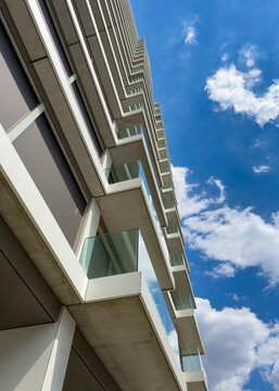 Modern apartment building balconies against blue sky. A low angle perspective shows rows of glass balconies on a high rise concrete building under scattered clouds.