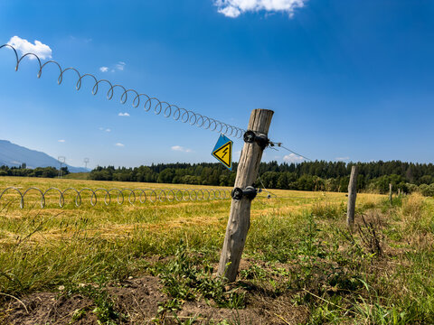 Electric fence line in countryside meadow. Spiral steel wire extends across dry yellow grass under a clear blue sky with distant mountain ranges.