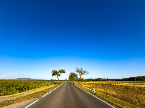 Wide asphalt road leading through rural fields under blue sky. A wide asphalt road extends straight toward distant hills between agricultural fields under a vast blue sky.