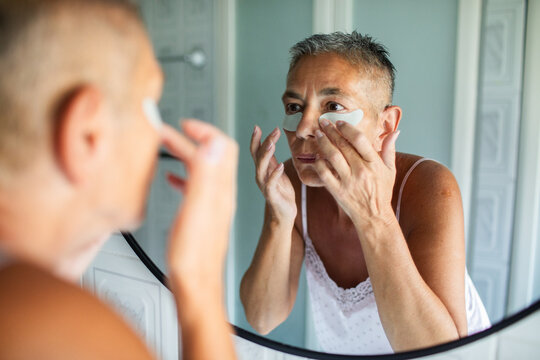 Senior woman applying eye patches in bathroom mirror