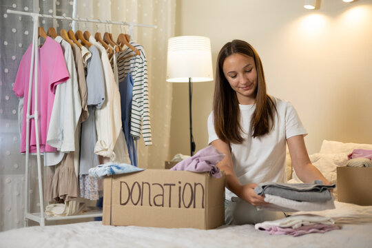 Teenage girl collecting and sorting her clothes for donation, placing garments into a cardboard box marked Donation.