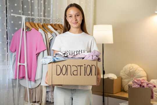 Teenage girl collecting and sorting her clothes for donation, placing garments into a cardboard box marked Donation.