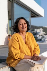 Naklejka premium Mature woman smiling with open book on sunlit terrace background in yellow blouse for lifestyle wallpaper