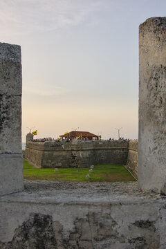 Vertical sunset shot framed through a fortress embrasure looking toward distant ramparts and outdoor cafes. Historical tourism and architectural framing concept.