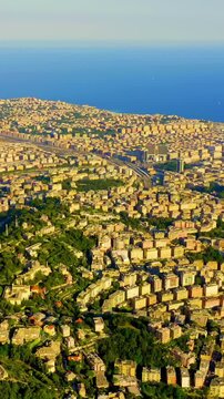 Vertical video. Genoa, Italy. Aerial panorama over the historic Porto Antico and dense terracotta rooftops, with cargo ships and cranes lining the deep blue Ligurian Sea under bright summer light. Dr