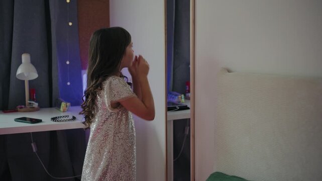Little girl wearing a sparkling sequin dress looks at her hairstyle in front of a mirror in her bedroom.