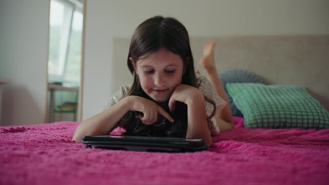 A young girl lies on a pink bed at home, happily playing games and tapping on a digital tablet screen.