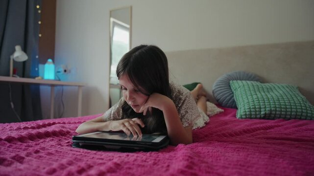 A young girl lies on a cozy bed in her bedroom while tapping and interacting with a digital tablet screen.