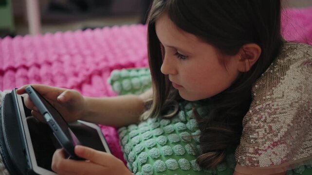 A focused young girl lies on a bed with a pink blanket, using a digital tablet at home.