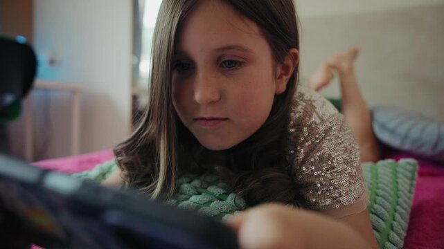 Little girl focused on playing a mobile game on her smartphone while lying down comfortably on a bed at home.
