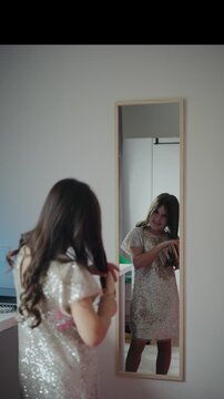 Vertical video of a girl in a silver sequin dress brushing her long brunette hair looking into a full-length wall mirror at home.