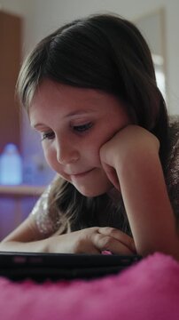 Vertical video of a focused young girl lying down in bed and using a digital tablet at home.
