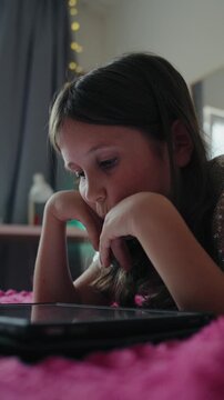 Vertical video of a focused little girl lying on a cozy bed and tapping on a digital tablet screen at home.