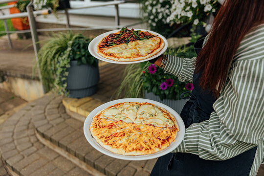 Restaurant worker carries two plated pizzas with assorted cheese, vegetable, corn, and sauce toppings through an outdoor dining area with flowers and greenery