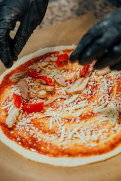 Black-gloved hands place red pepper pieces onto uncooked pizza topped with sliced chicken, shredded cheese, and tomato sauce during preparation
